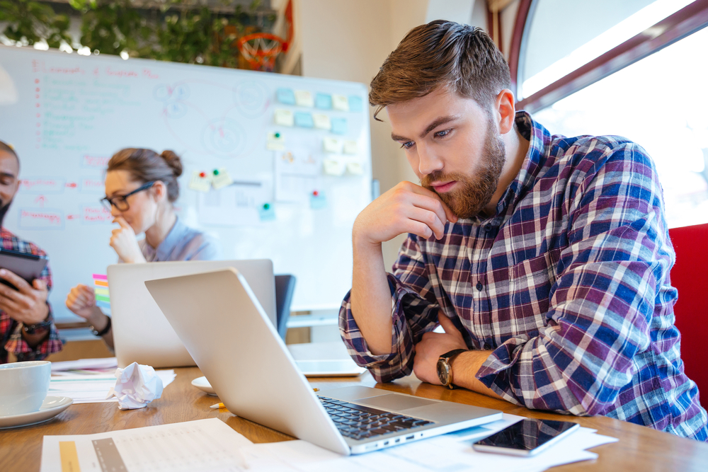Concentrated bearded young man using laptop while his friends studying together Concentrated bearded young man using laptop while his friends studying together